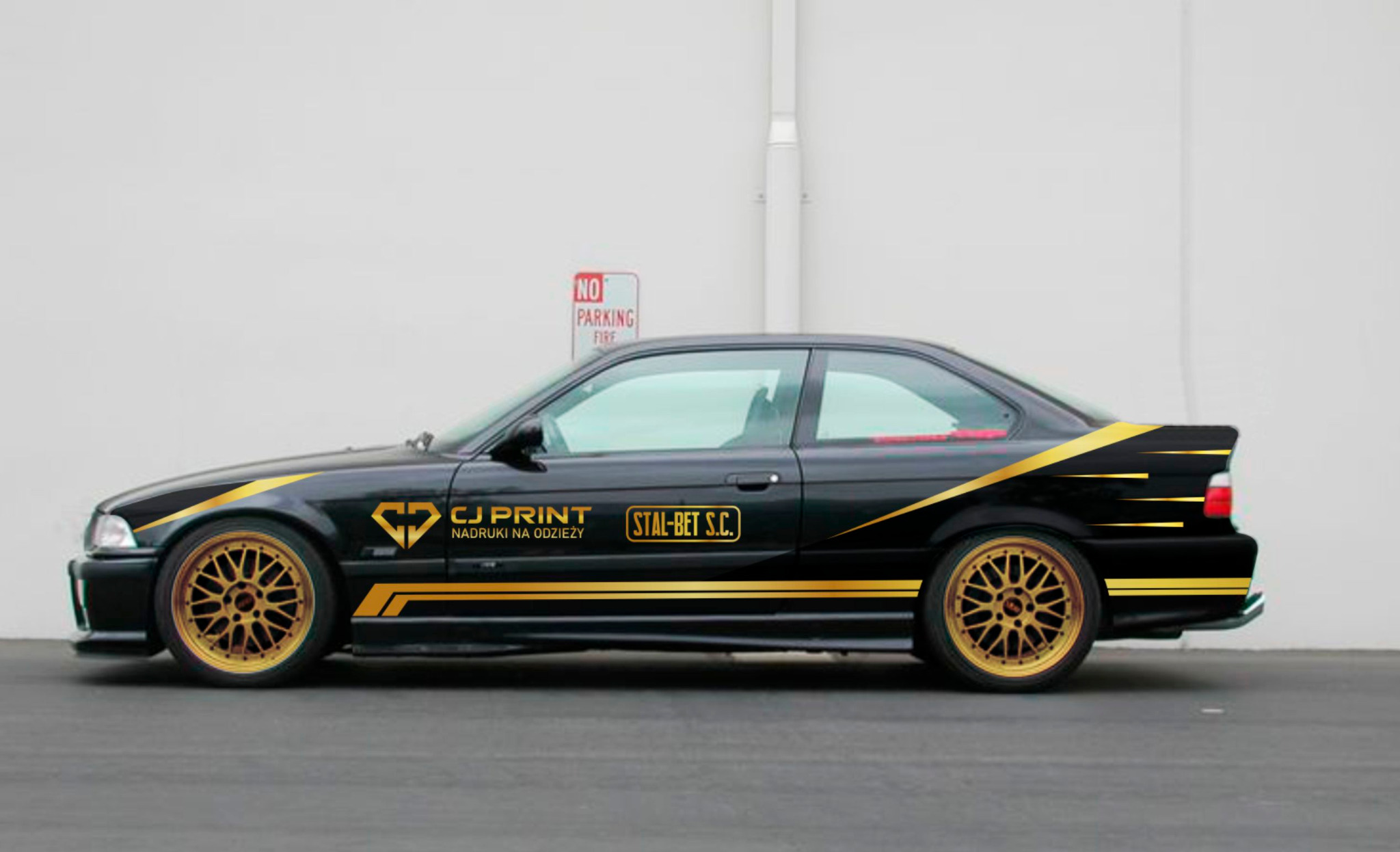 Czarny samochód BMW E36 z złotymi felgami i naklejonymi złotymi paskami oraz logo firm CJ Print i Stal-Bet S.C. na białym tle ściany.