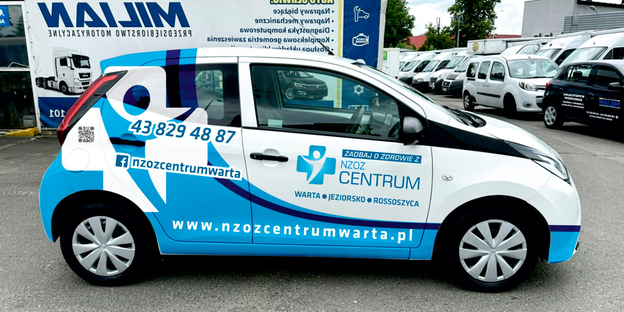 Samochód oklejony folią z niebiesko-białą grafiką i danymi kontaktowymi NZOZ Centrum, zaparkowany na parkingu z innymi pojazdami w tle.