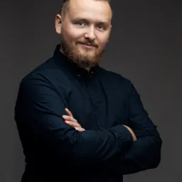 Portret brodatego mężczyzny w ciemnej koszuli, z założonymi rękami na szarym tle, patrzącego w obiektyw z lekkim uśmiechem.