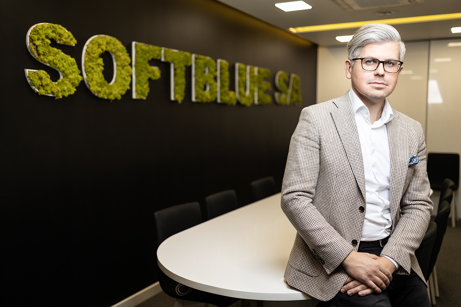 Portret biznesowy mężczyzny w okularach i tweedowej marynarce, opierającego się o biały stół konferencyjny na tle czarnej ściany z logo firmy wykonanym z mchu.