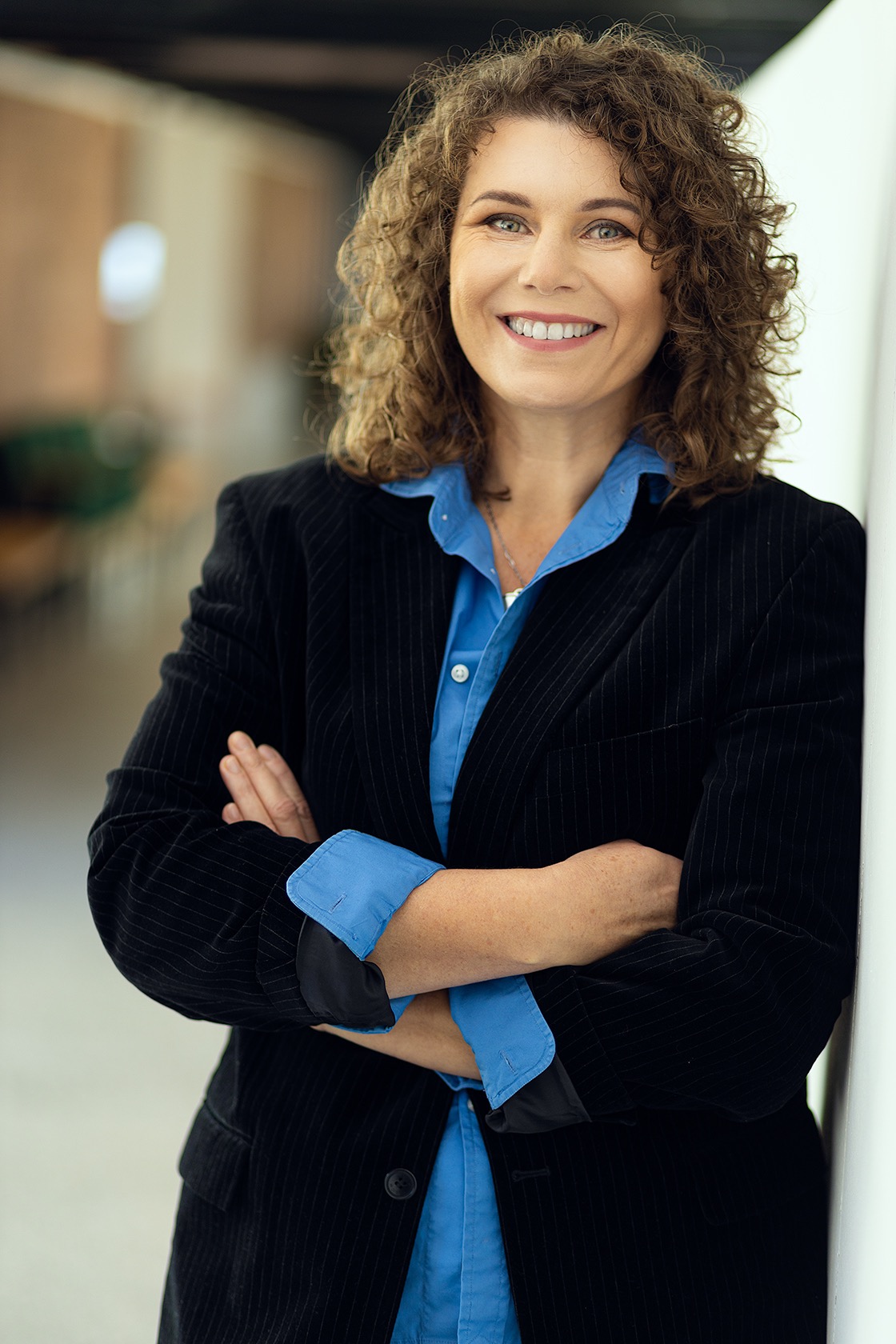 Uśmiechnięta kobieta w czarnym żakiecie i niebieskiej koszuli, oparta o białą ścianę, z rękami skrzyżowanymi na piersiach.