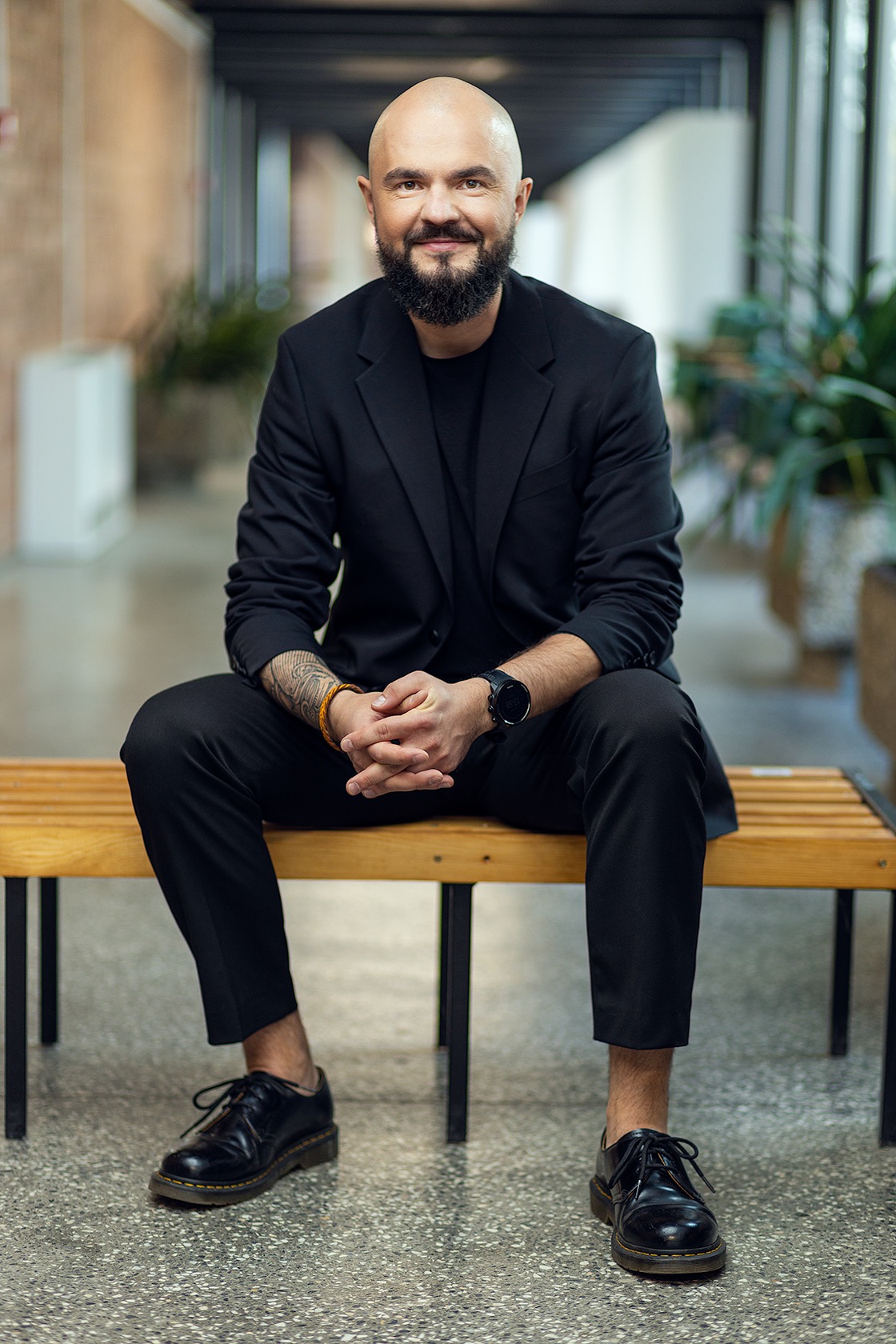 Portret mężczyzny z brodą, ubranego w czarny garnitur, siedzącego na drewnianej ławce w nowoczesnym wnętrzu z roślinami.