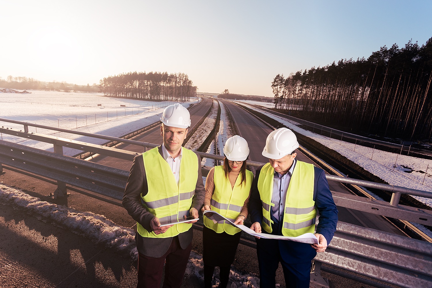 Trzech inżynierów w kaskach i kamizelkach odblaskowych analizuje plan na wiadukcie drogi ekspresowej w zimowej scenerii, widoczna zasnieżona łąka i las w tle.
