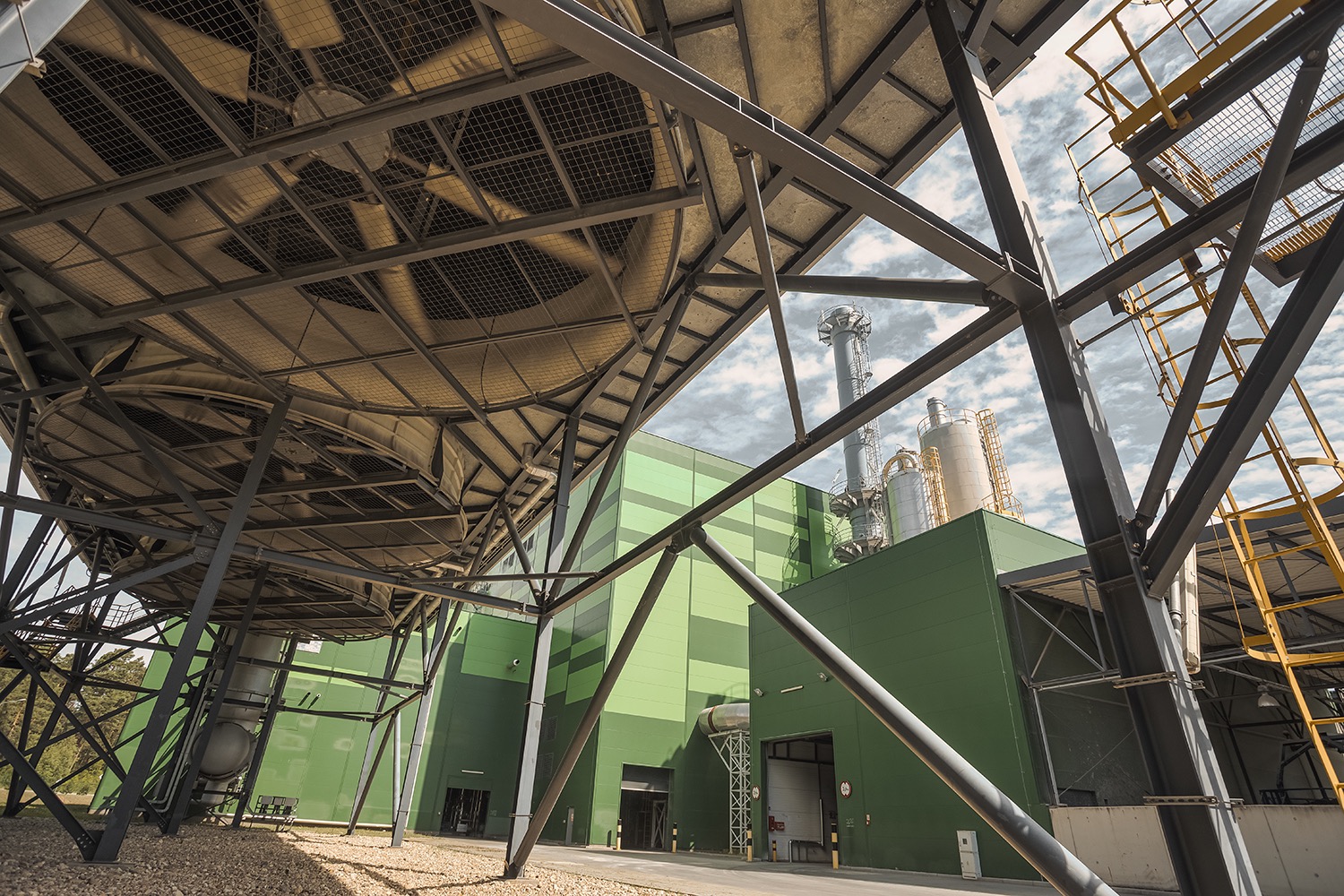 Widok z dołu na wentylator chłodniczy zamontowany na konstrukcji stalowej, z zielonym budynkiem przemysłowym i kominami w tle.