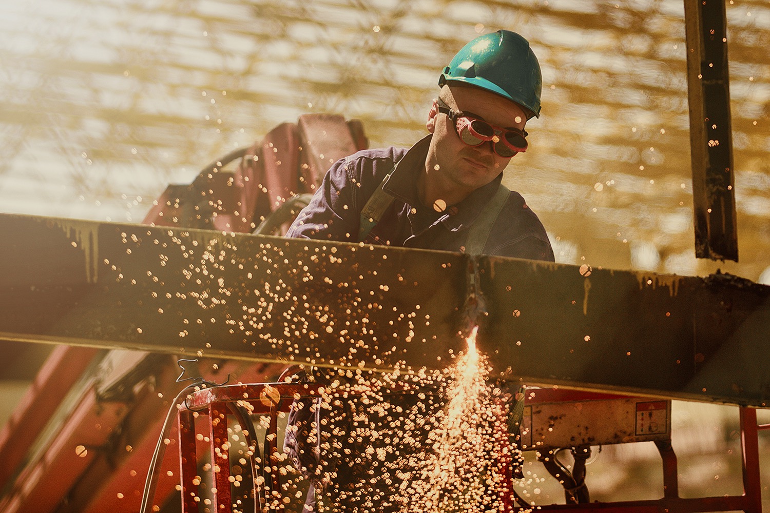Pracownik w kasku i okularach ochronnych spawa metalową konstrukcję na wysokości, iskry rozbryzgują się w powietrzu.