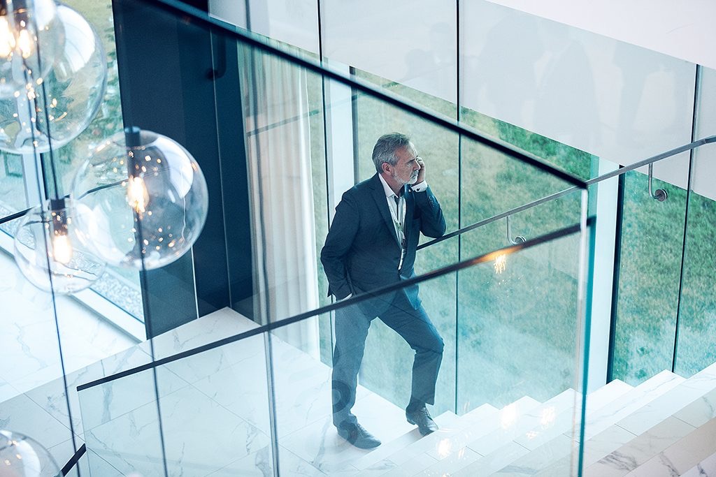 Mężczyzna w garniturze rozmawia przez telefon, wchodząc po marmurowych schodach z przeszkloną balustradą w nowoczesnym wnętrzu z widokiem na zieleń przez okno.