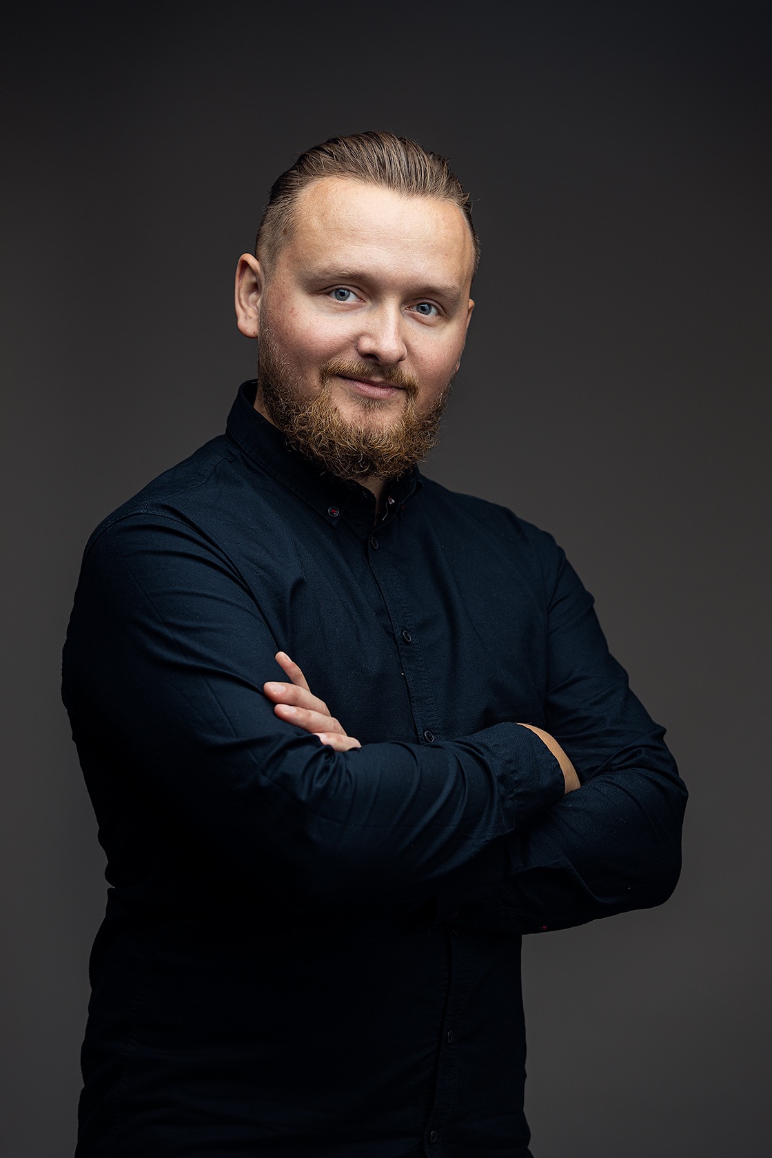 Portret brodatego mężczyzny w ciemnej koszuli, z założonymi rękami na szarym tle, patrzącego w obiektyw z lekkim uśmiechem.