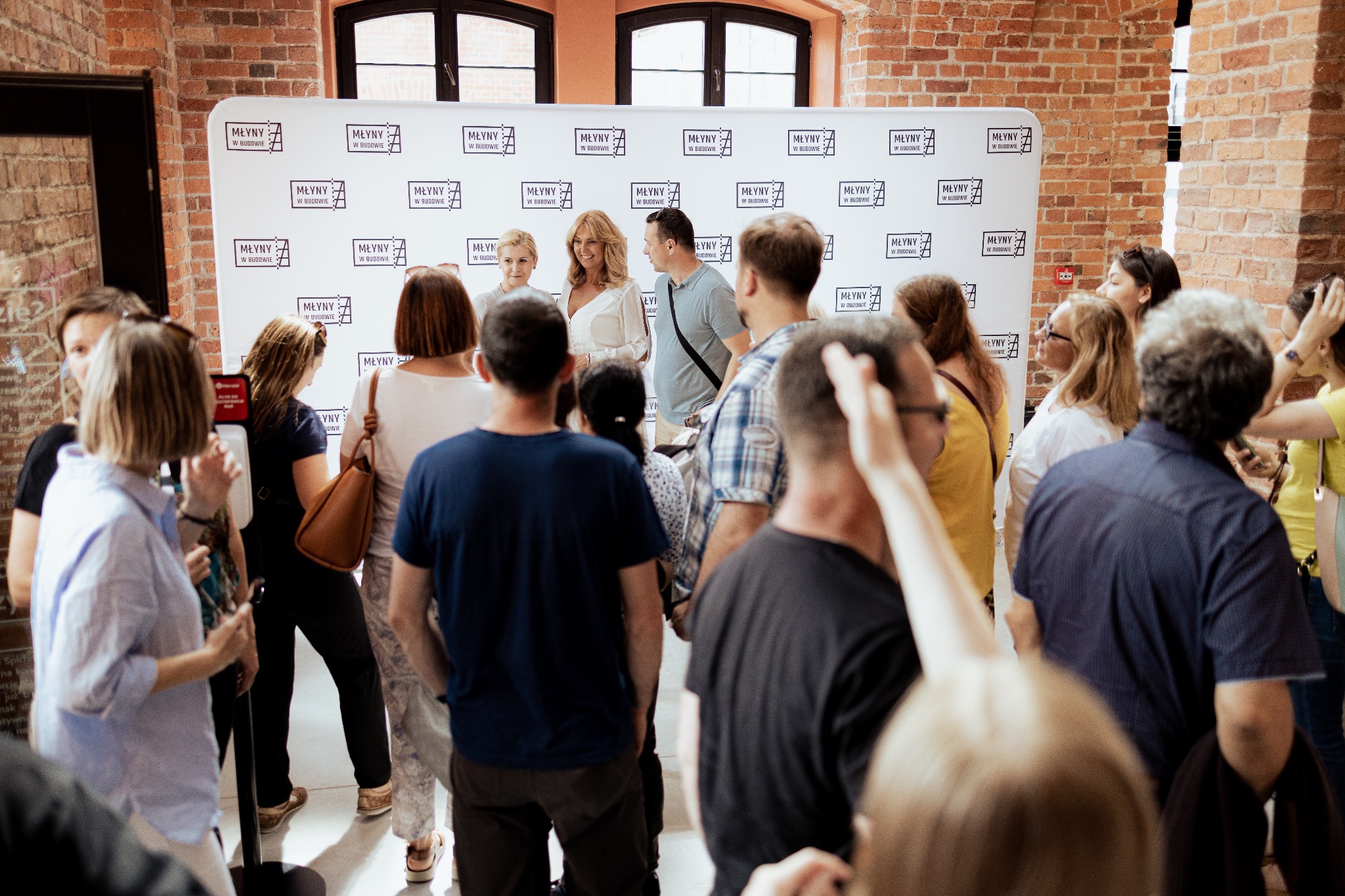 Dwie kobiety w białych sukienkach pozują na tle ściany z logo 'Młyny w Budowie', otoczone przez grupę ludzi, w ceglanym wnętrzu.