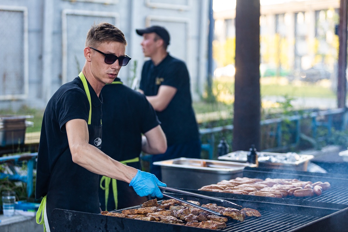 Młody mężczyzna w okularach słonecznych i niebieskich rękawiczkach obraca mięso na grillu, w tle widoczni inni mężczyźni i budynki.