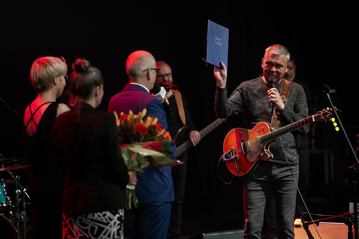 Muzyk z gitarą na scenie, trzyma w ręku dyplom, obok osoby z bukietem kwiatów i mikrofonem, w tle zespół muzyczny.