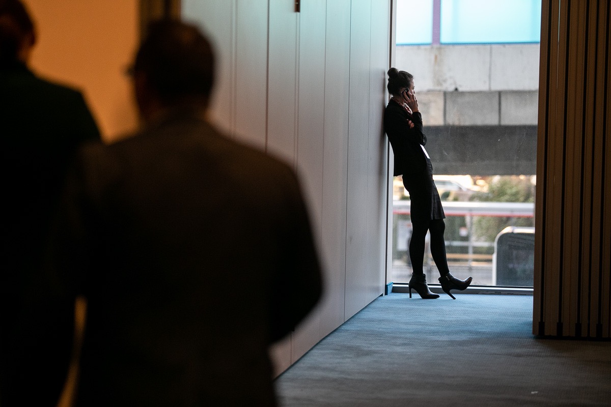 Kobieta w czarnym garniturze opiera się o ścianę przy oknie, rozmawiając przez telefon; w tle zamazane sylwetki dwóch mężczyzn.