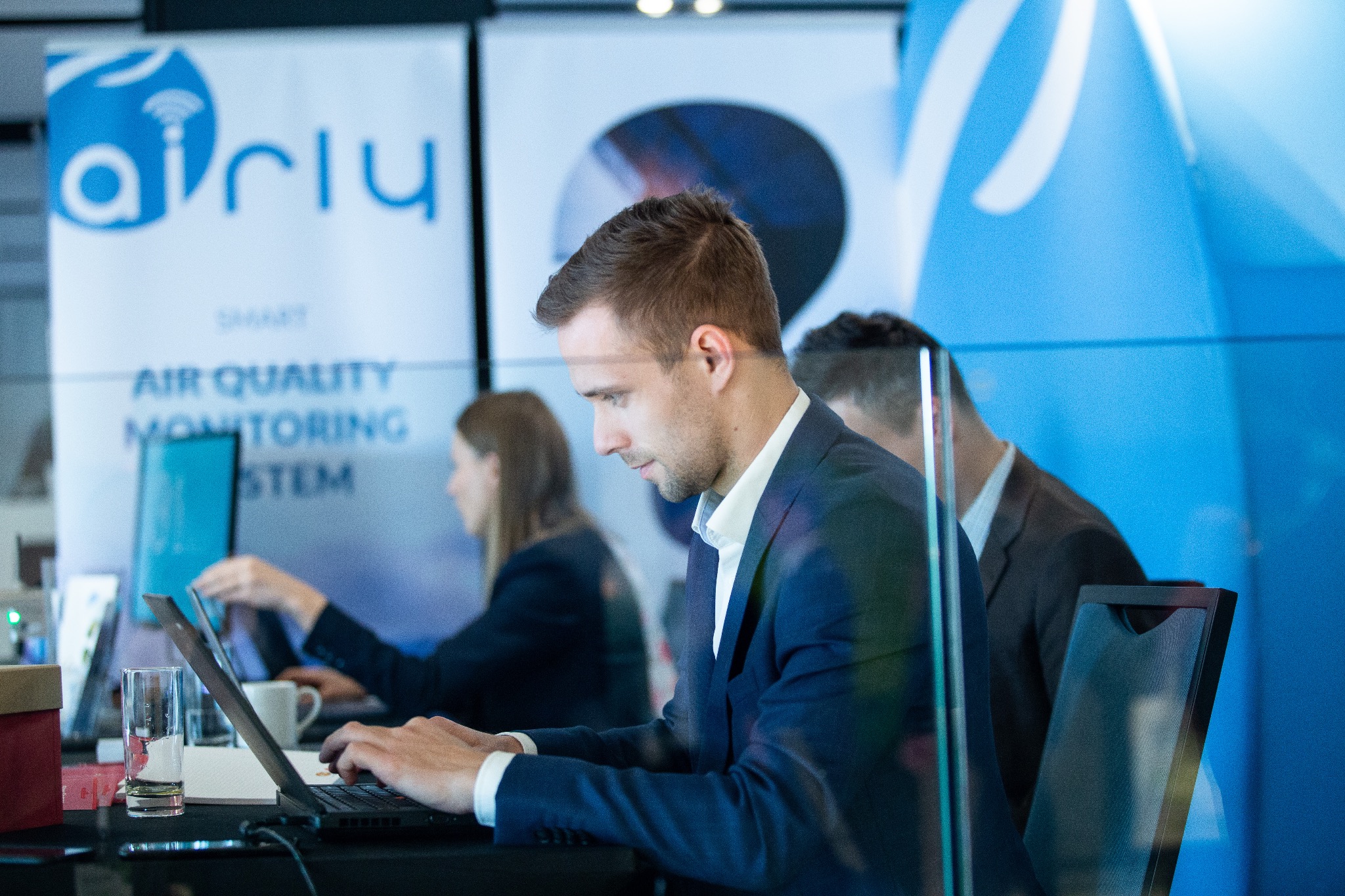 Mężczyzna w garniturze pisze na laptopie na tle stoiska z logo firmy Airly promującej system monitoringu jakości powietrza, widoczna szklanka wody i filiżanka kawy.