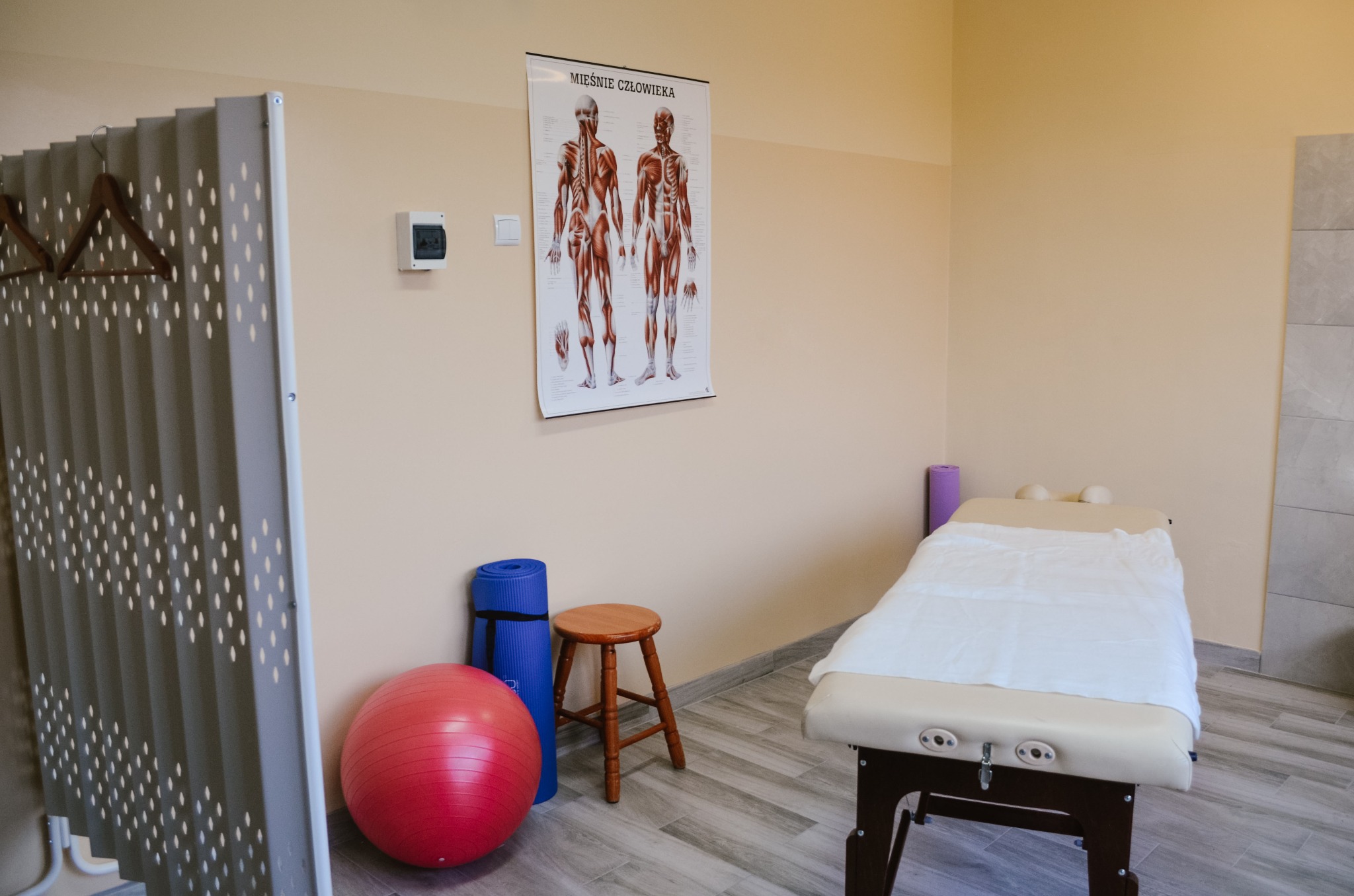 Jasny gabinet z leżanką przykrytą białym prześcieradłem, plakatem z układem mięśniowym człowieka na ścianie, piłką do ćwiczeń, matą do jogi i drewnianym stołkiem.