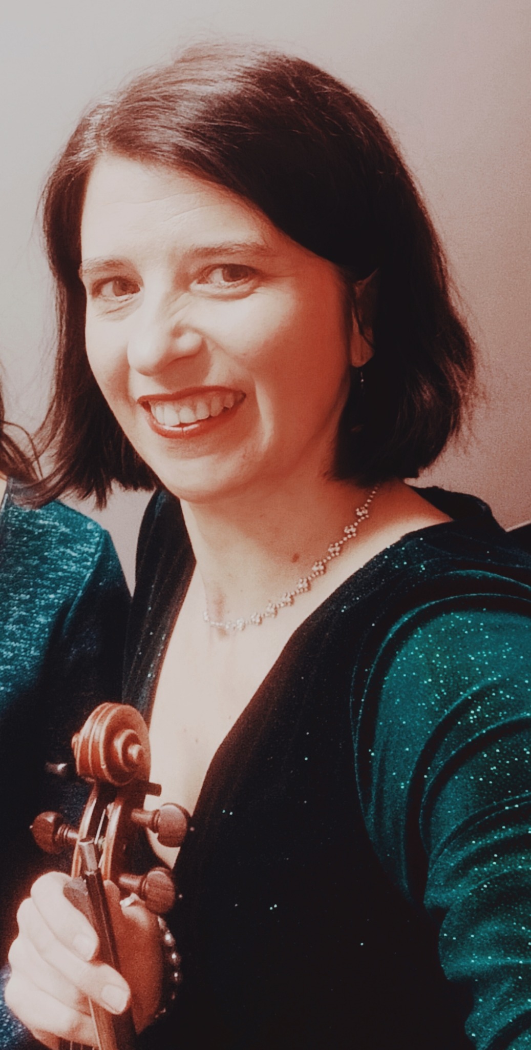 Uśmiechnięta kobieta w błyszczącej, zielonej sukience trzymająca skrzypce, widoczny fragment drugiej osoby po lewej stronie.