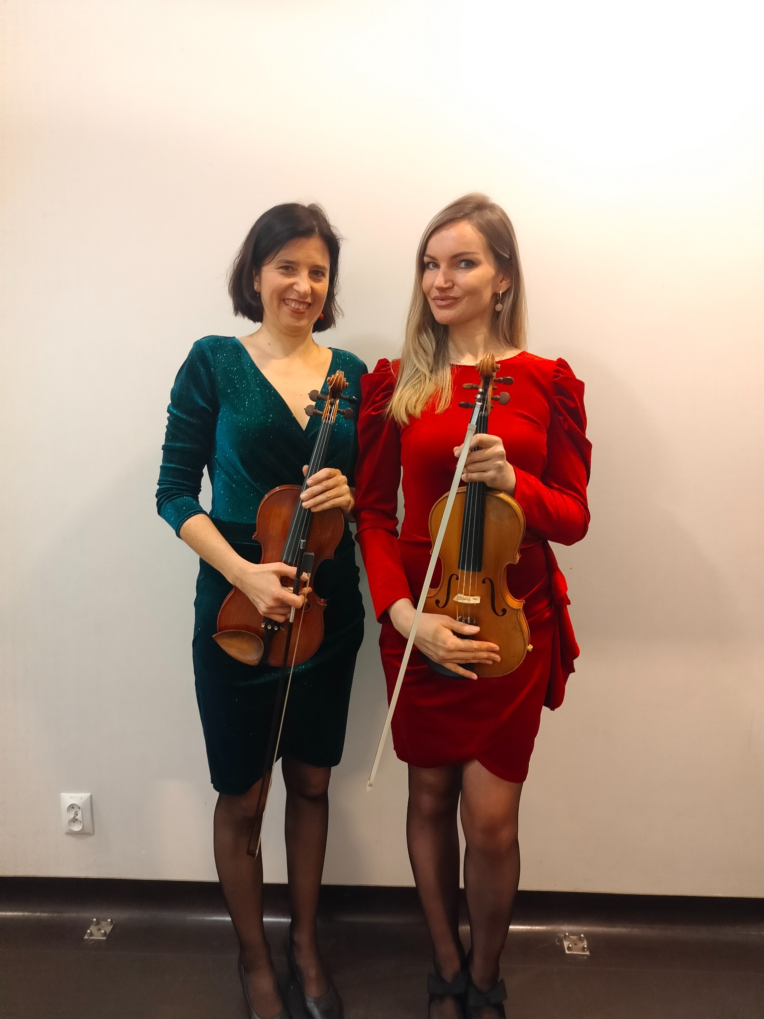 Dwie uśmiechnięte kobiety w eleganckich sukienkach, trzymające skrzypce i smyczki na tle białej ściany. Jedna w szmaragdowej, druga w czerwonej sukience.