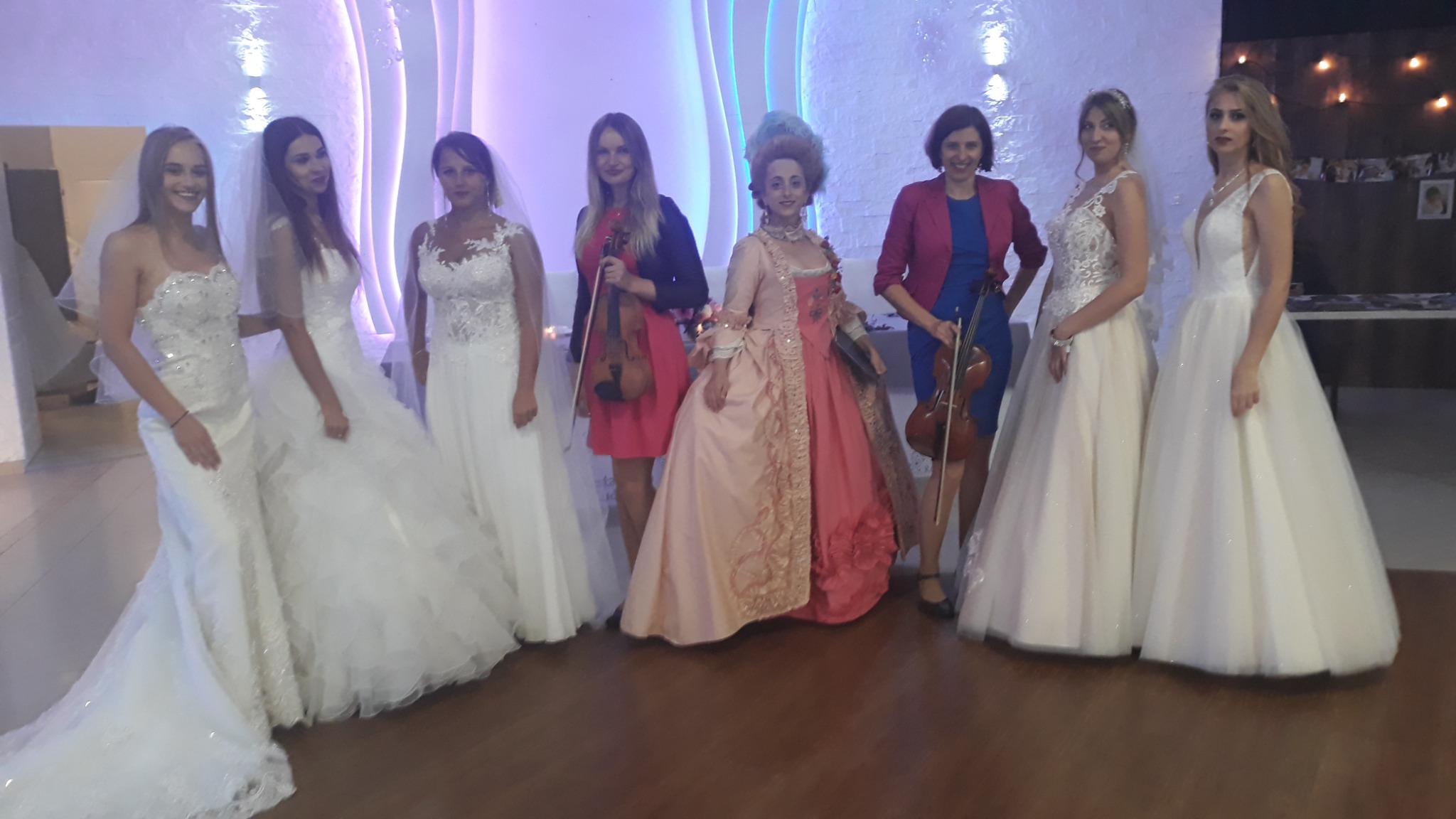 Kobiety w sukniach ślubnych i historycznej, niektóre trzymają skrzypce, prezentacja na tle dekoracji świetlnych.