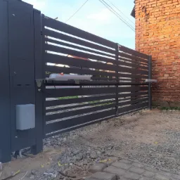 Nowoczesna, grafitowa brama przesuwna z automatyką, widoczne siłowniki i skrzynka sterownicza, na tle ceglanego budynku i gruntu z kamieniami.