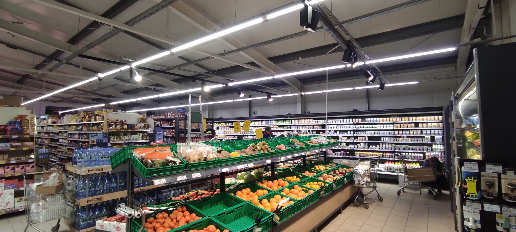 Wnętrze sklepu spożywczego z widocznymi półkami pełnymi produktów, oświetleniem sufitowym i klientem w tle, z bliska ujęte kosze z owocami i warzywami.