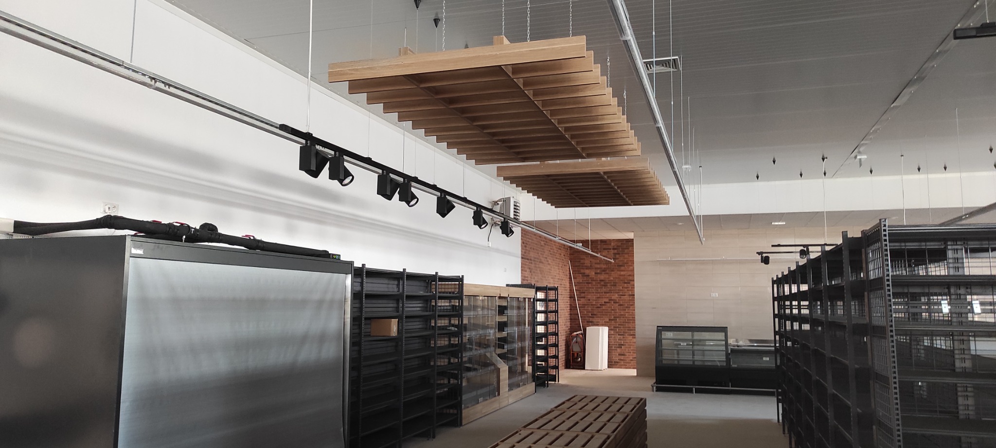 Wnętrze sklepu w trakcie urządzania: widoczne regały, oświetlenie szynowe oraz drewniane panele sufitowe. Instalacja elektryczna widoczna na górze chłodziarki. Część regałów już zamontowana.
