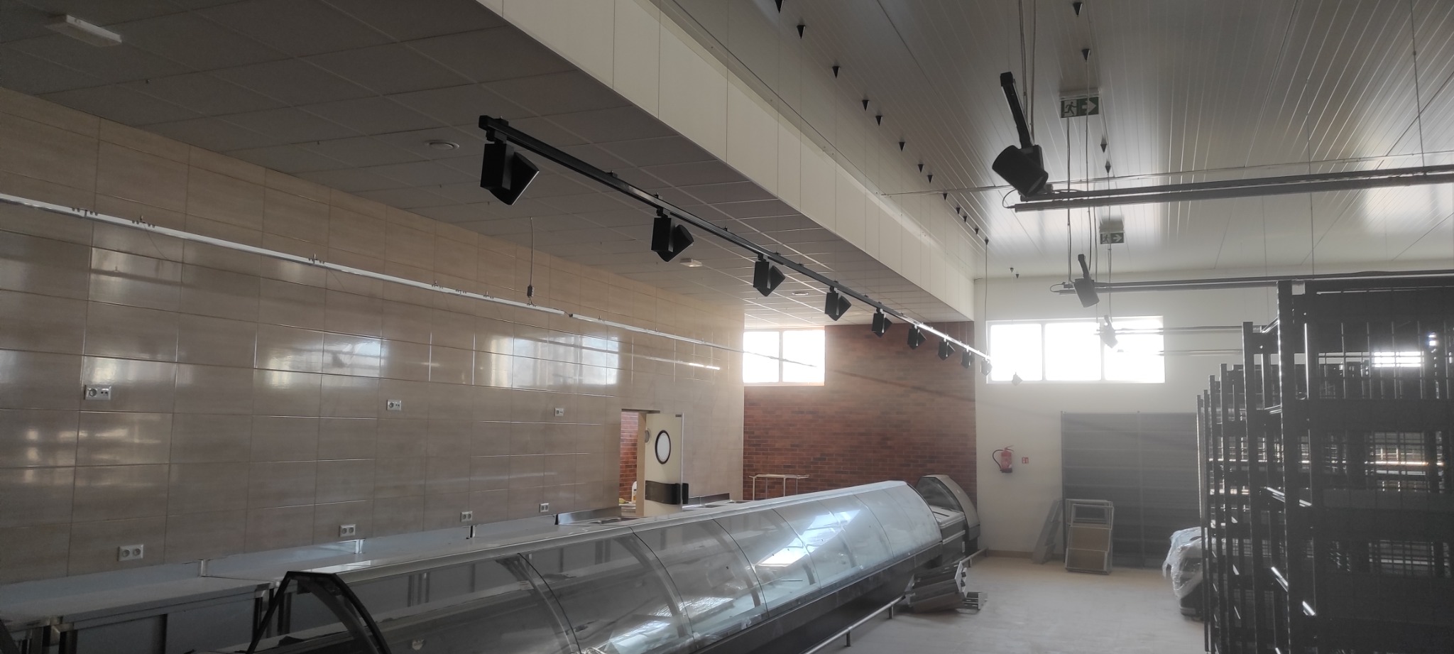 Wnętrze sklepu z widocznymi instalacjami elektrycznymi: szyny z reflektorami sufitowymi, gniazdka elektryczne na ścianach wyłożonych kafelkami, oraz ekspozytory chłodnicze przed ceglaną ścianą.