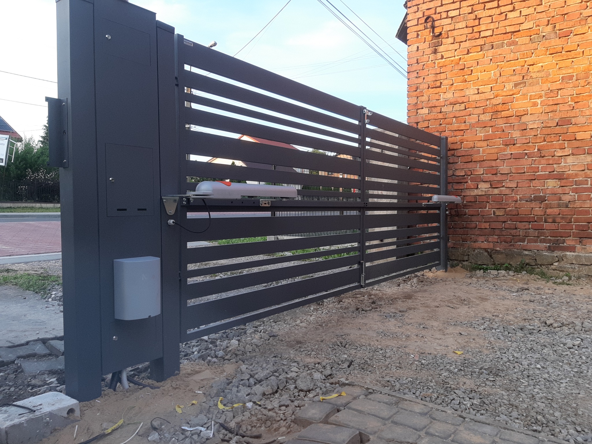Nowoczesna, grafitowa brama przesuwna z automatyką, widoczne siłowniki i skrzynka sterownicza, na tle ceglanego budynku i gruntu z kamieniami.