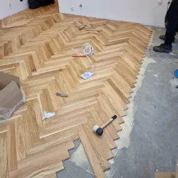 Proces układania parkietu w jodełkę francuską, widoczne drewniane klepki, klej, gumowy młotek, narzędzia i częściowo ułożona podłoga w pomieszczeniu w trakcie remontu.