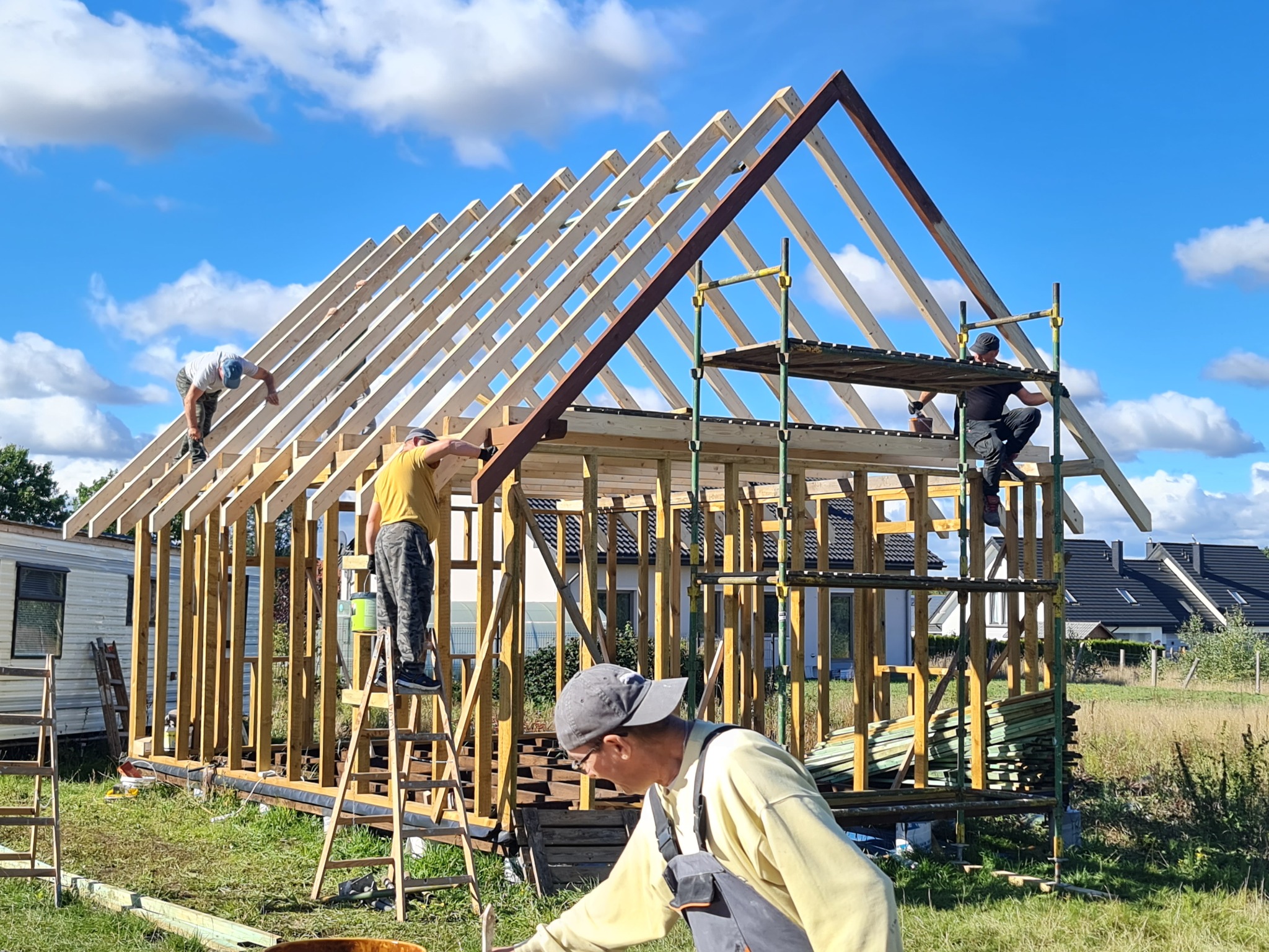 Montaż szkieletu drewnianego domu jednorodzinnego: robotnicy na rusztowaniach i drabinach mocują drewniane elementy konstrukcji dachu, w tle błękitne niebo z chmurami i zabudowa jednorodzinna.