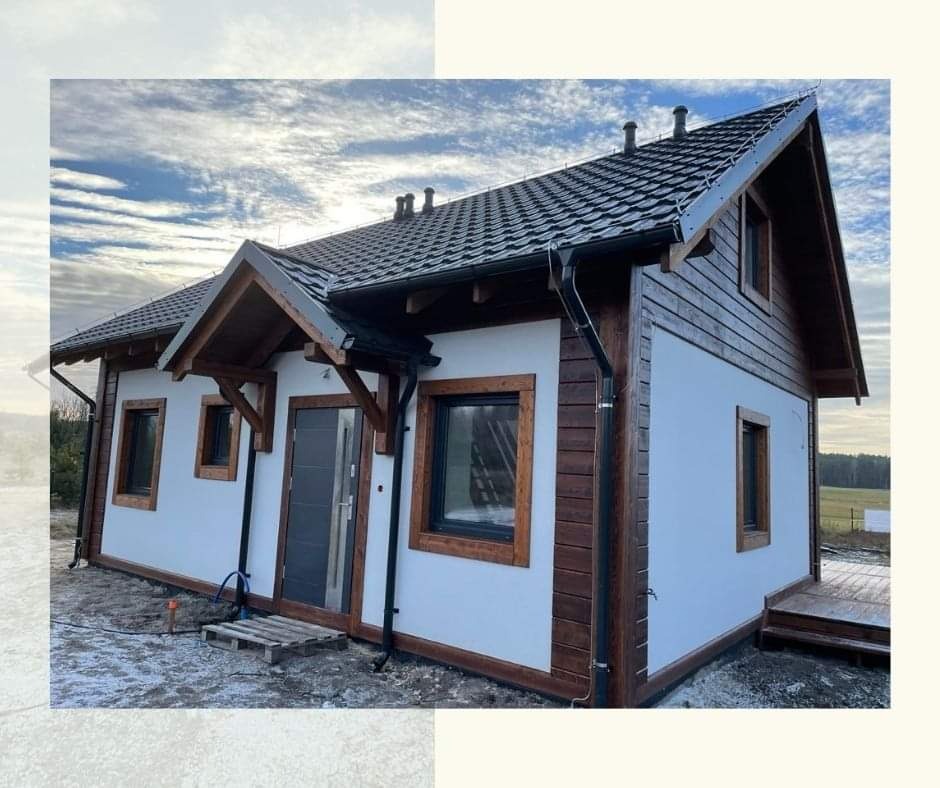 Gotowy dom szkieletowy z białymi ścianami i ciemnobrązowymi elementami drewnianymi, dach pokryty czarną dachówką, widoczne rynny i okna z ciemnymi ramami.