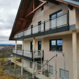 Nowoczesny dom z dwoma balkonami z metalowymi balustradami o poziomym układzie prętów, widoczne rusztowania sugerują prace wykończeniowe.