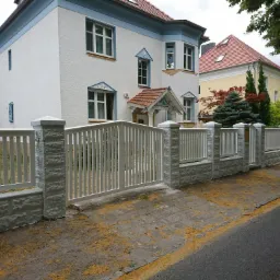 Biała brama wjazdowa z pionowymi szczeblami i betonowymi słupkami o fakturze cegły, prowadząca do posesji z dwoma budynkami mieszkalnymi w tle.