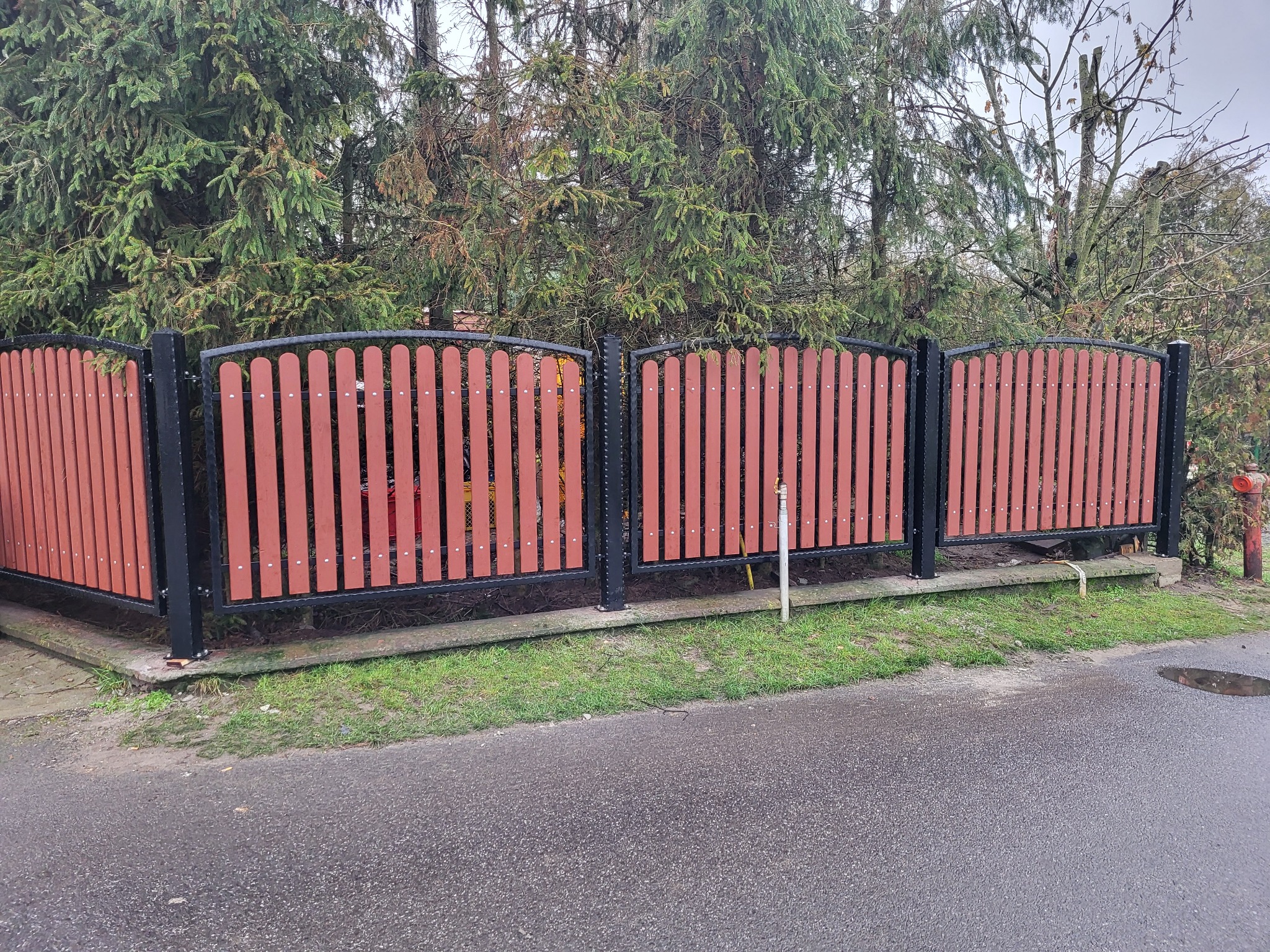 Fragment ogrodzenia z drewnianymi sztachetami w kolorze terakoty i czarną metalową ramą, widoczne z poziomu ulicy w pochmurny dzień.