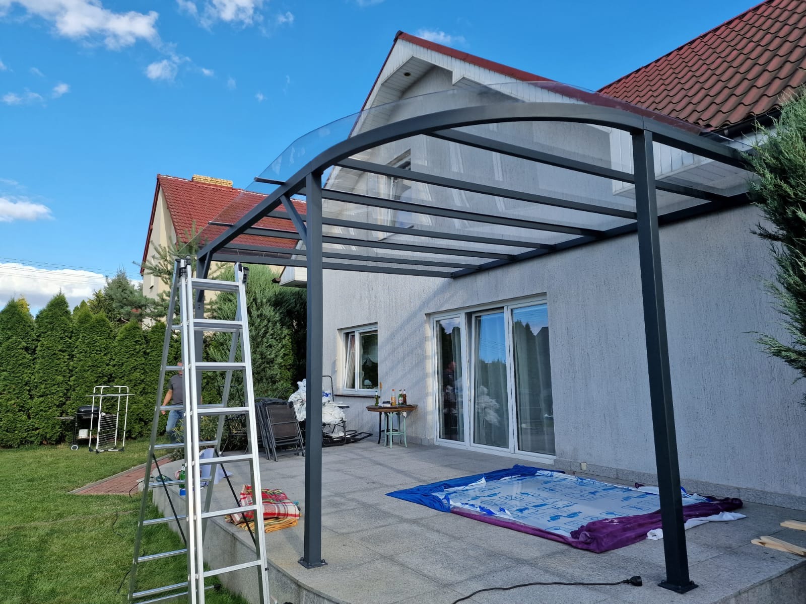Zadaszenie tarasu z metalową konstrukcją i szklanym dachem w trakcie montażu, widoczna drabina i zabezpieczona podłoga, dom jednorodzinny w tle.
