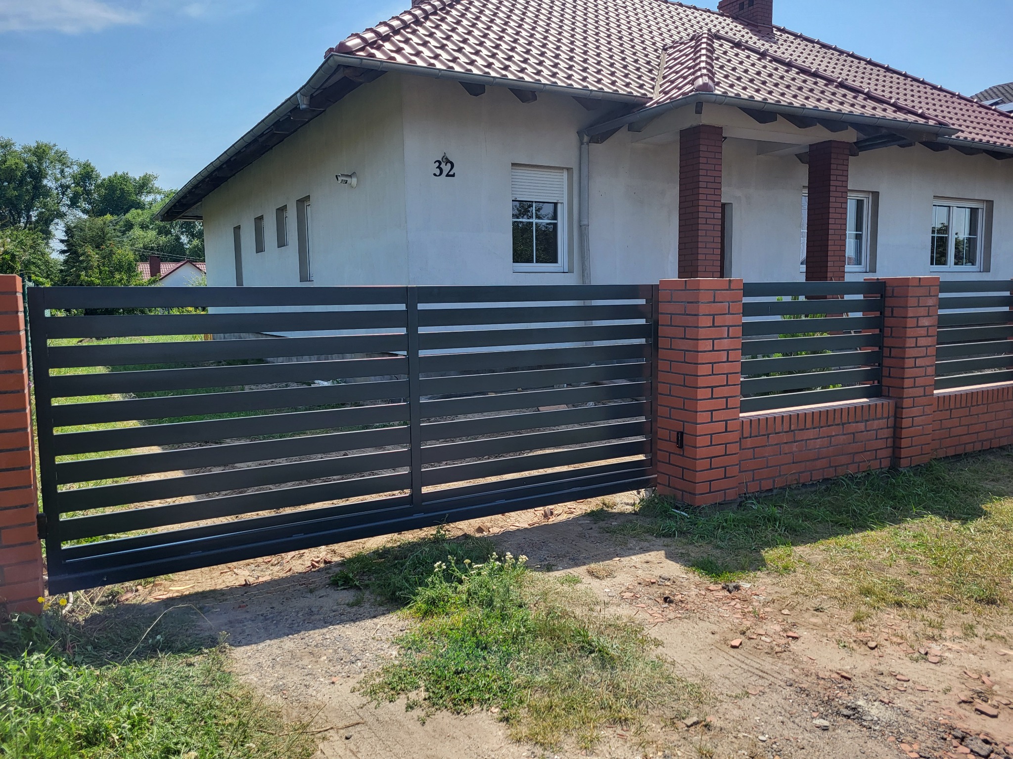 Nowoczesna brama wjazdowa z poziomych, grafitowych paneli, osadzona na ceglanych słupkach, przed domem z jasną elewacją i numerem 32.