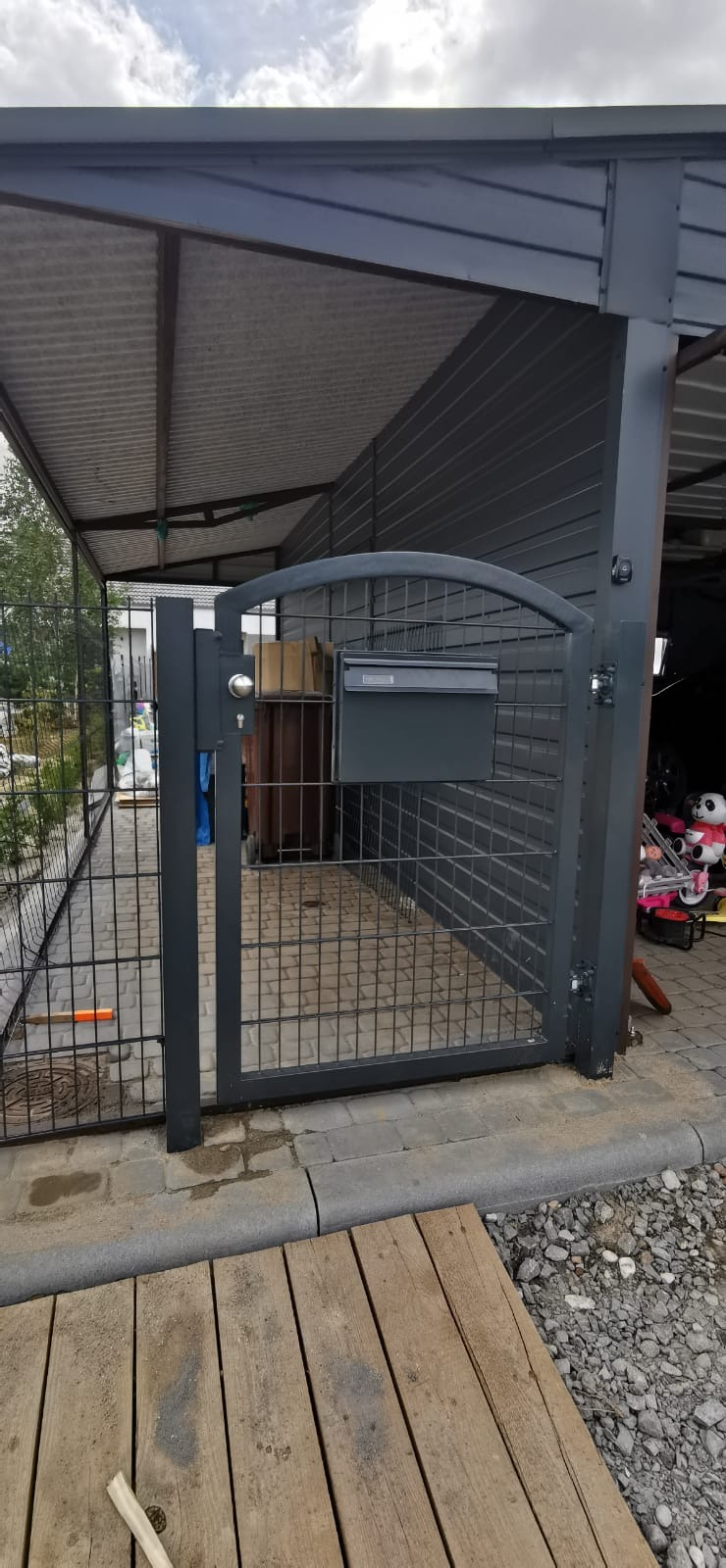Szara, metalowa furtka z zamontowaną skrzynką na listy, część ogrodzenia panelowego, widok z zewnątrz na podwórko z kostką brukową i elementami drewnianymi.