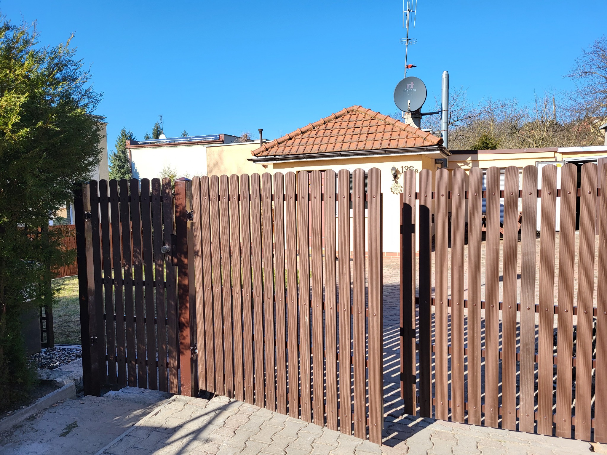 Brunatna, drewnopodobna brama wjazdowa z pionowych desek, prowadząca do domu z czerwoną dachówką i anteną satelitarną na tle błękitnego nieba.