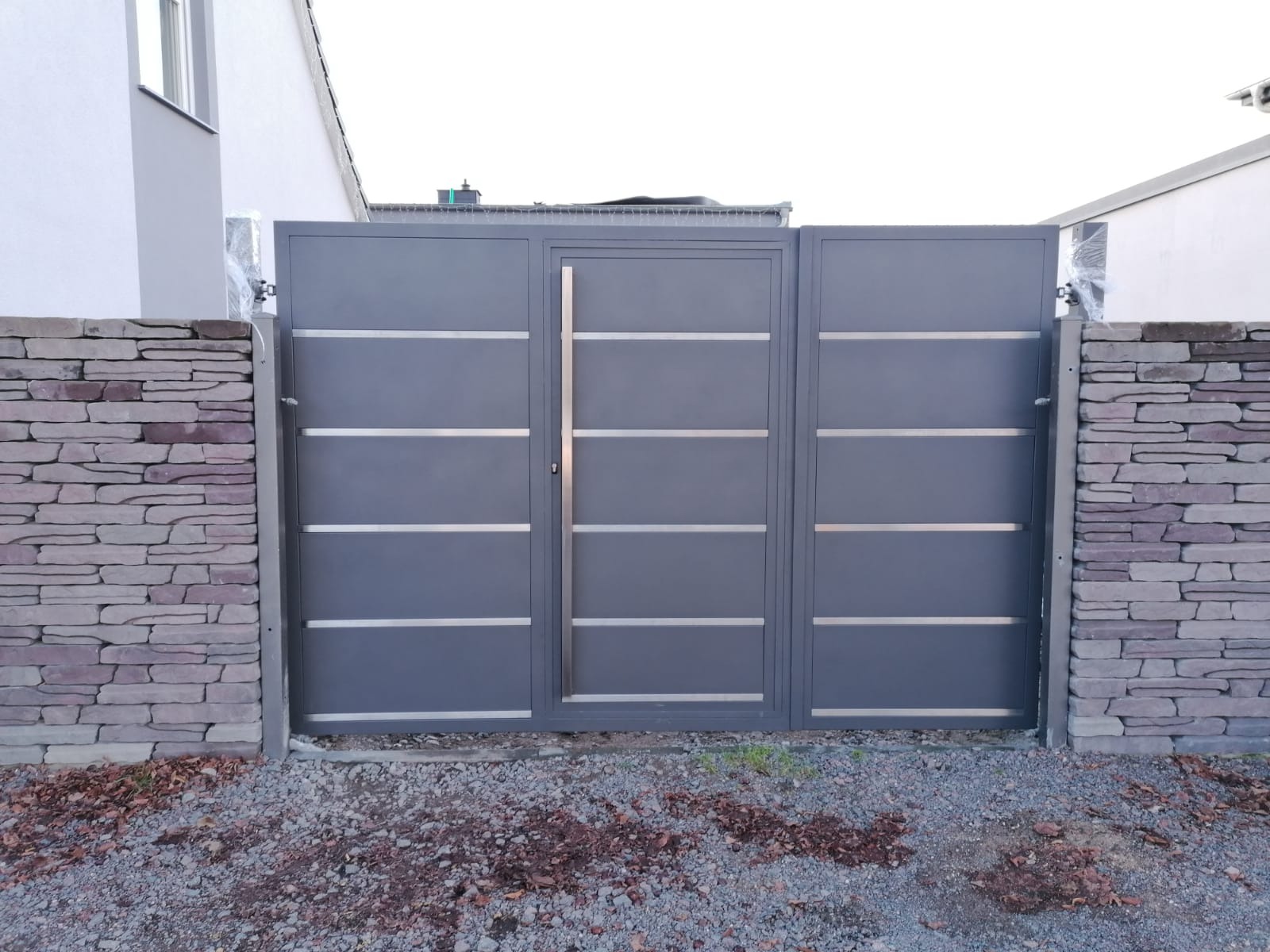 Nowoczesna, szara brama wjazdowa z poziomymi, srebrnymi akcentami, zamontowana w Gorzowie Wielkopolskim. Brama otoczona kamiennym murem, z widocznymi elementami automatyki w folii ochronnej.