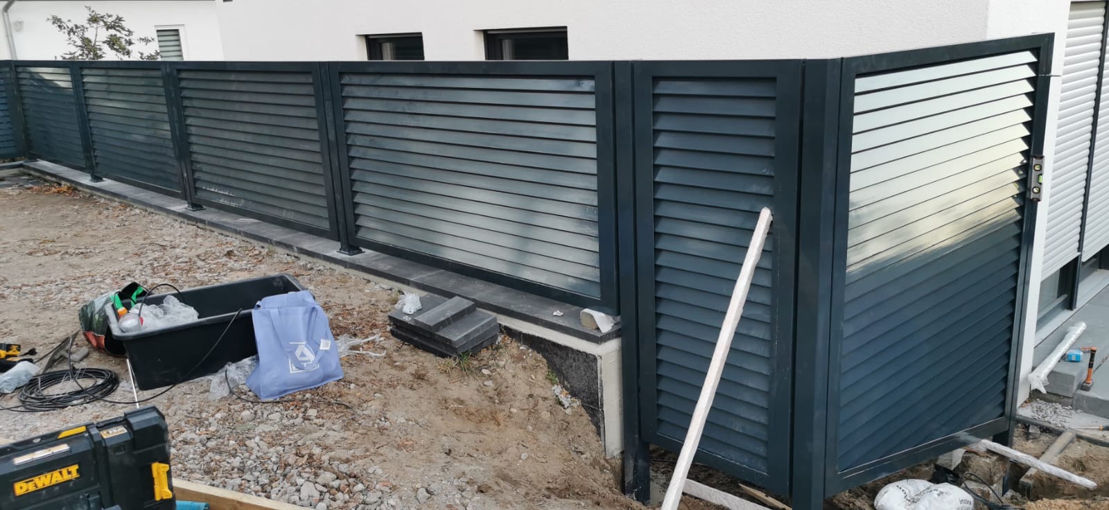 Nowoczesne ogrodzenie z poziomych, metalowych lameli w kolorze grafitowym, widoczne narzędzia i materiały budowlane na ziemi, sugerujące montaż w toku.