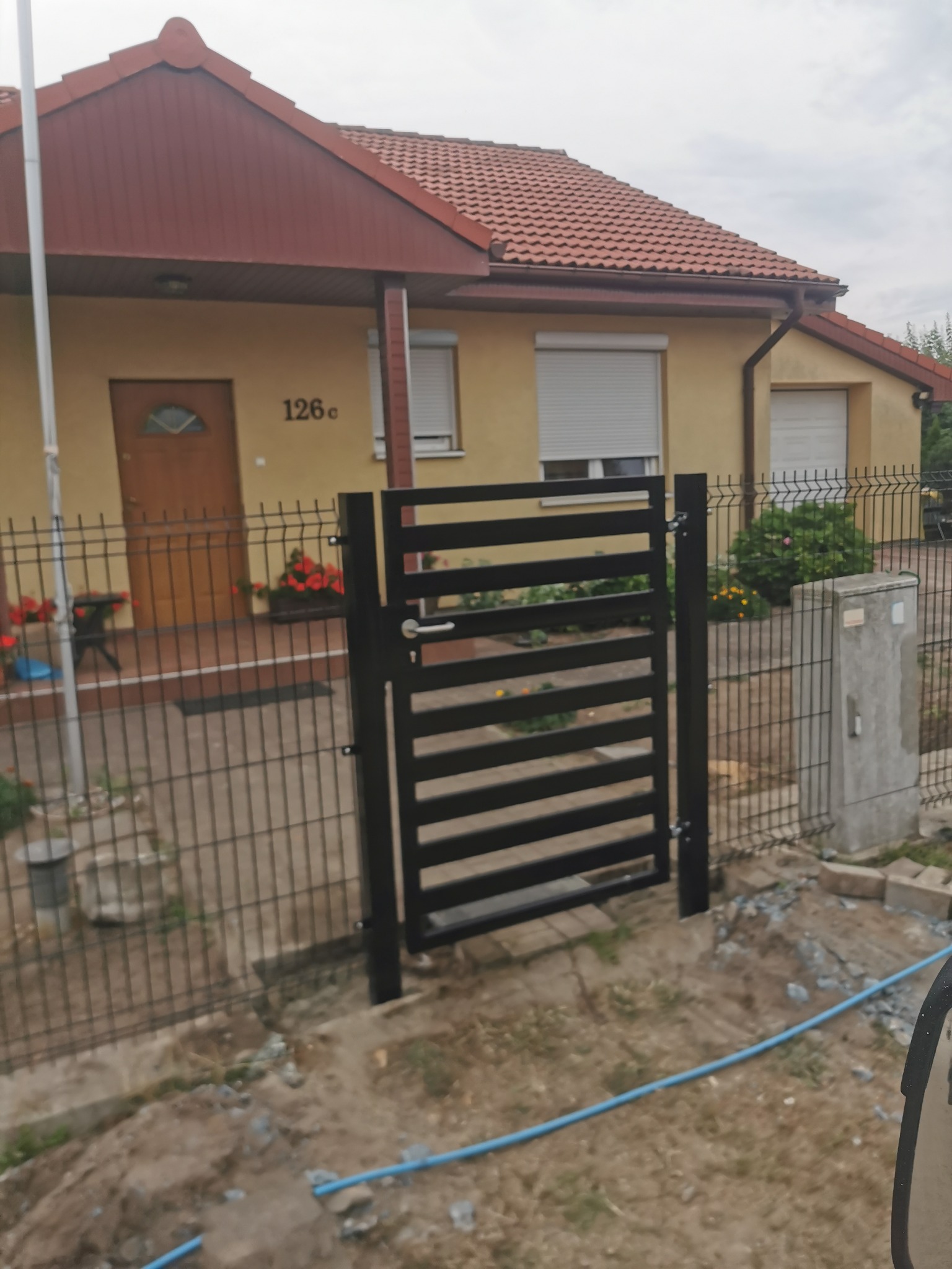 Nowoczesna, czarna furtka z poziomych profili, zamontowana w ogrodzeniu panelowym przed domem z żółtą elewacją i czerwonym dachem, widoczne świeże prace ziemne.