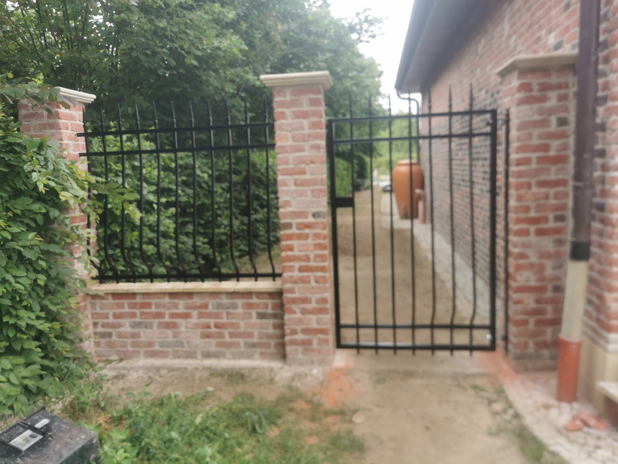 Czarne, metalowe ogrodzenie z bramką, osadzone na niskim murku z czerwonej cegły z białymi kapitelami, widok na zieleń ogrodu i fragment ceglanego budynku w tle.