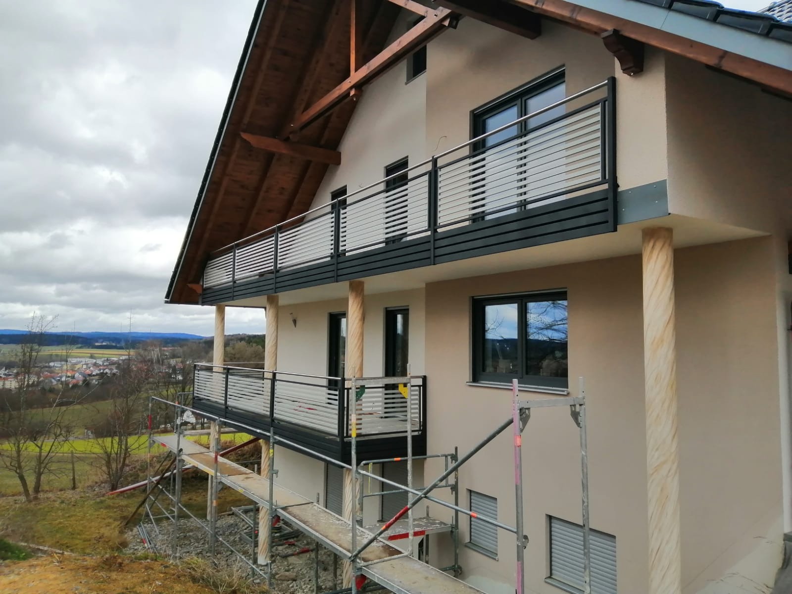 Nowoczesny dom z dwoma balkonami z metalowymi balustradami o poziomym układzie prętów, widoczne rusztowania sugerują prace wykończeniowe.