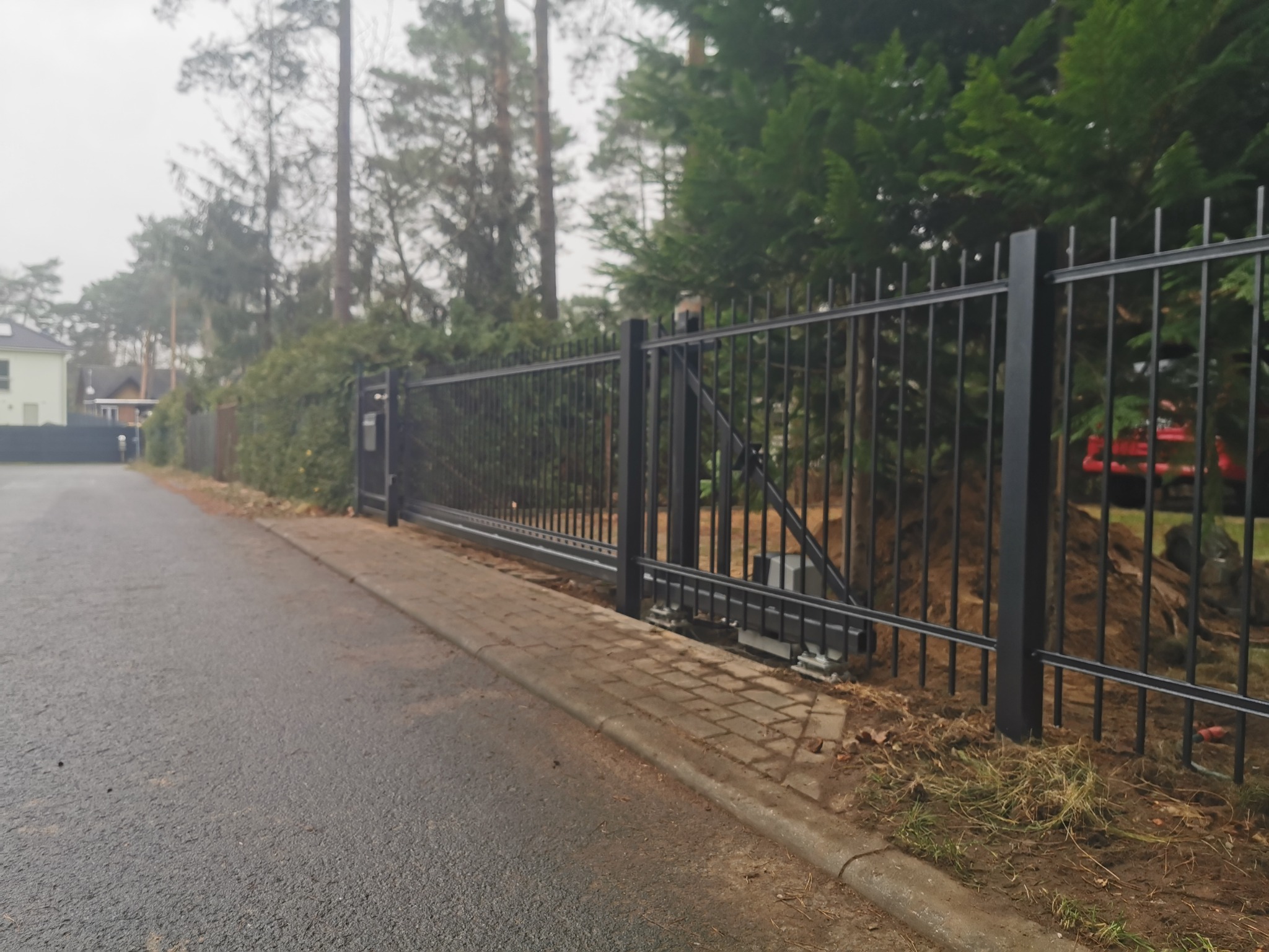 Automatyczna, przesuwna brama wjazdowa z czarnego metalu, widoczny mechanizm napędowy, kostka brukowa, drzewa w tle.