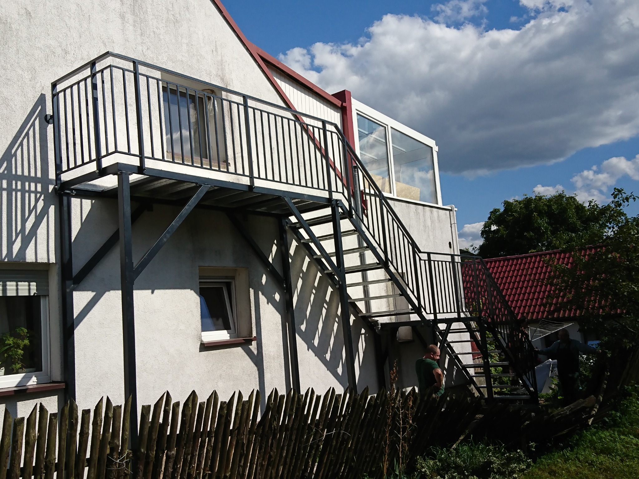 Zewnętrzne metalowe schody z balustradą, prowadzące do balkonu przy budynku mieszkalnym z białym tynkiem, widoczne zza drewnianego płotu.