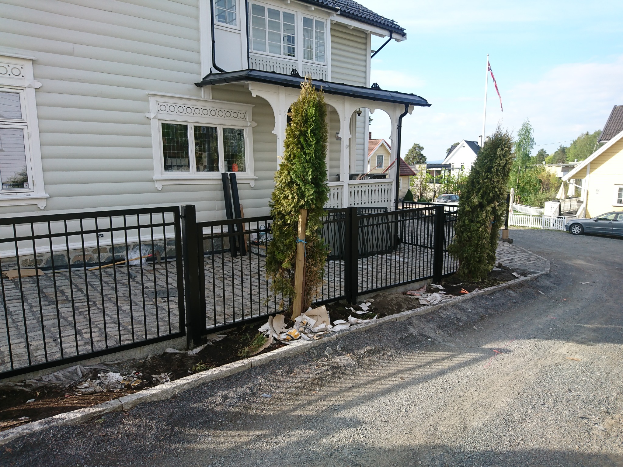 Czarny, metalowy płot palisadowy przed domem z jasną elewacją, świeżo posadzone tuje, gruz i resztki materiałów budowlanych wzdłuż krawężnika.