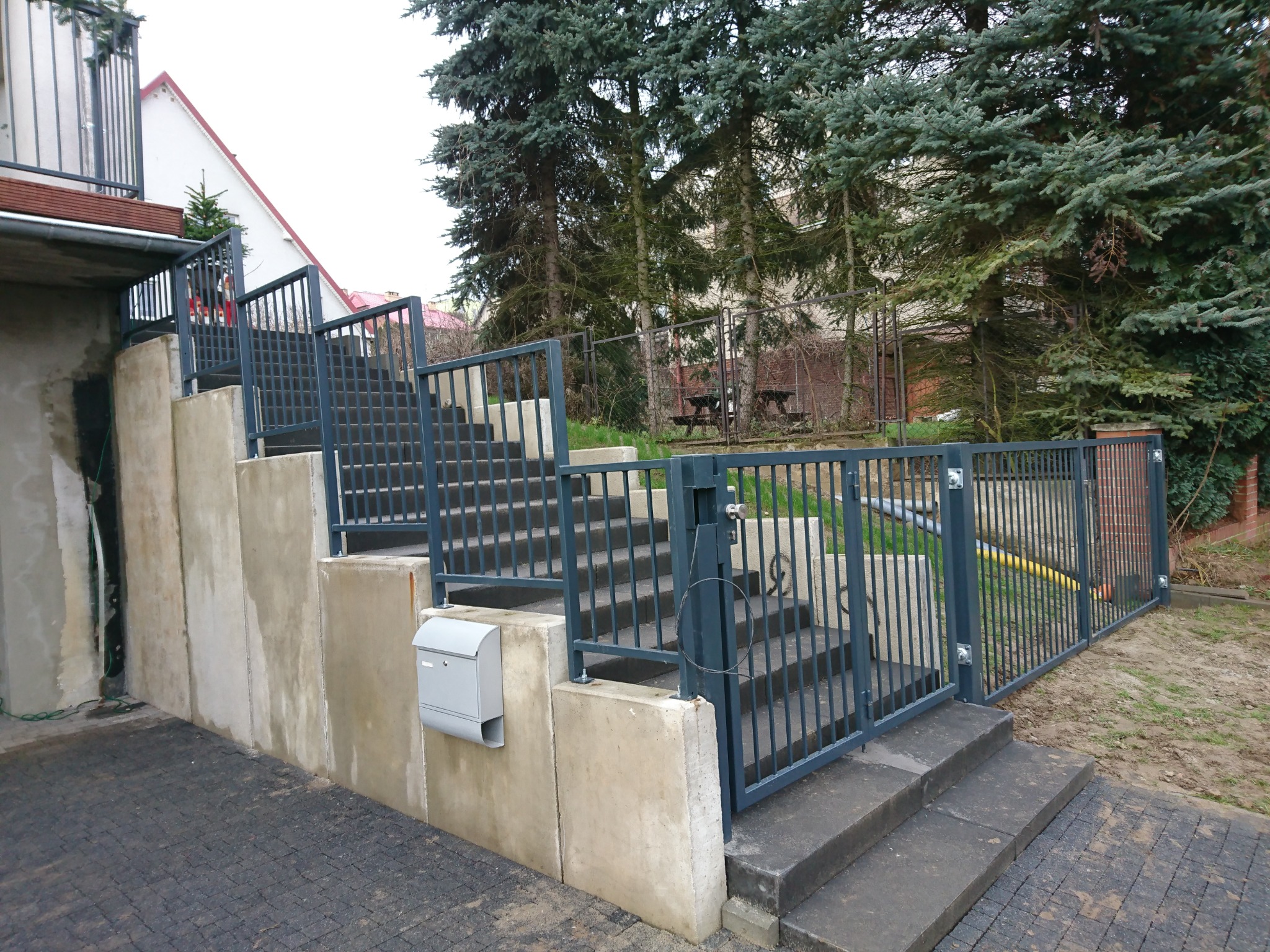 Zewnętrzne schody betonowe z metalową balustradą i furtką, szara skrzynka pocztowa na ścianie, widok na ogród z drzewami iglastymi.