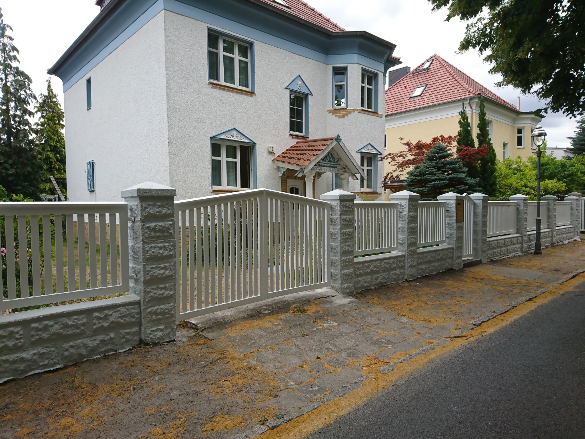 Biała brama wjazdowa z pionowymi szczeblami i betonowymi słupkami o fakturze cegły, prowadząca do posesji z dwoma budynkami mieszkalnymi w tle.