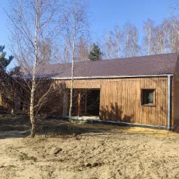 Nowoczesny, parterowy dom modułowy z drewnianą elewacją i brązowym dachem, otoczony piaszczystym terenem i młodymi brzozami, widziany w słoneczny dzień.