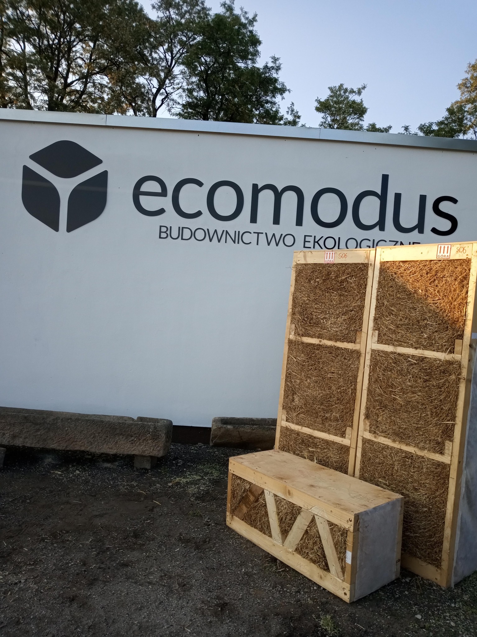 Dwa panele słomiane w drewnianej ramie, obok drewniana skrzynia wypełniona słomą, na tle białej ściany z logo firmy Ecomodus Budownictwo Ekologiczne, widoczne fragmenty kamiennych koryt.
