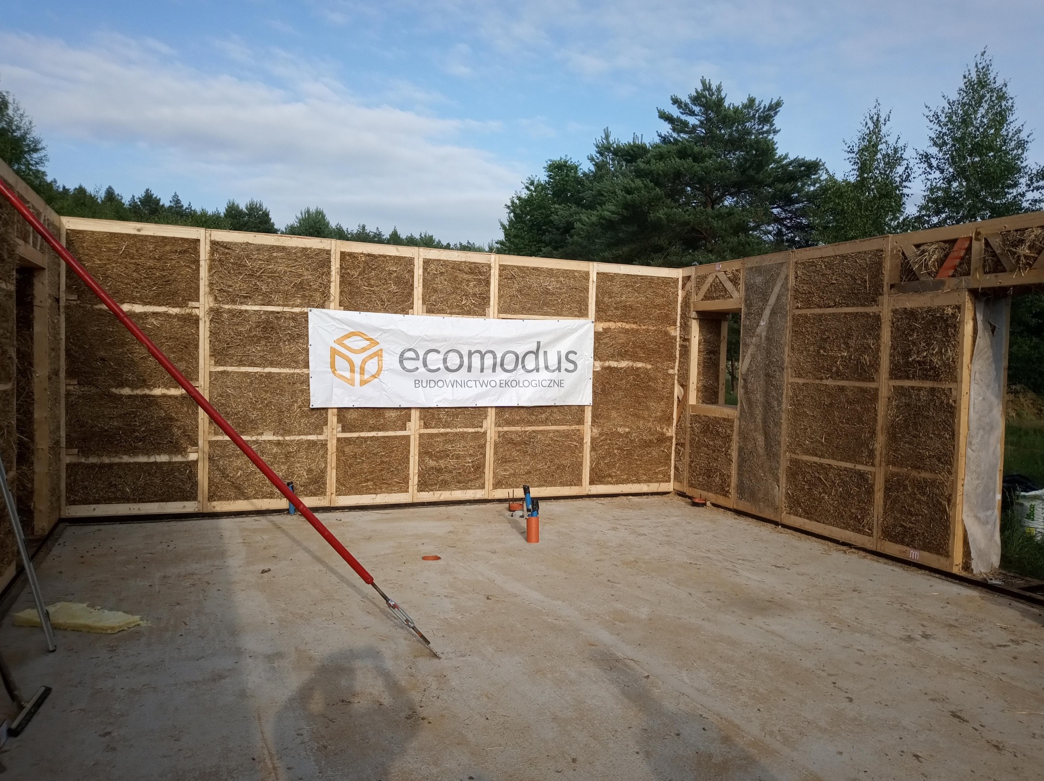 Wnętrze domu w trakcie budowy z bali słomianych, widoczna konstrukcja drewniana wypełniona słomą, baner firmy Ecomodus Budownictwo Ekologiczne na ścianie, betonowa podłoga w stanie surowym...