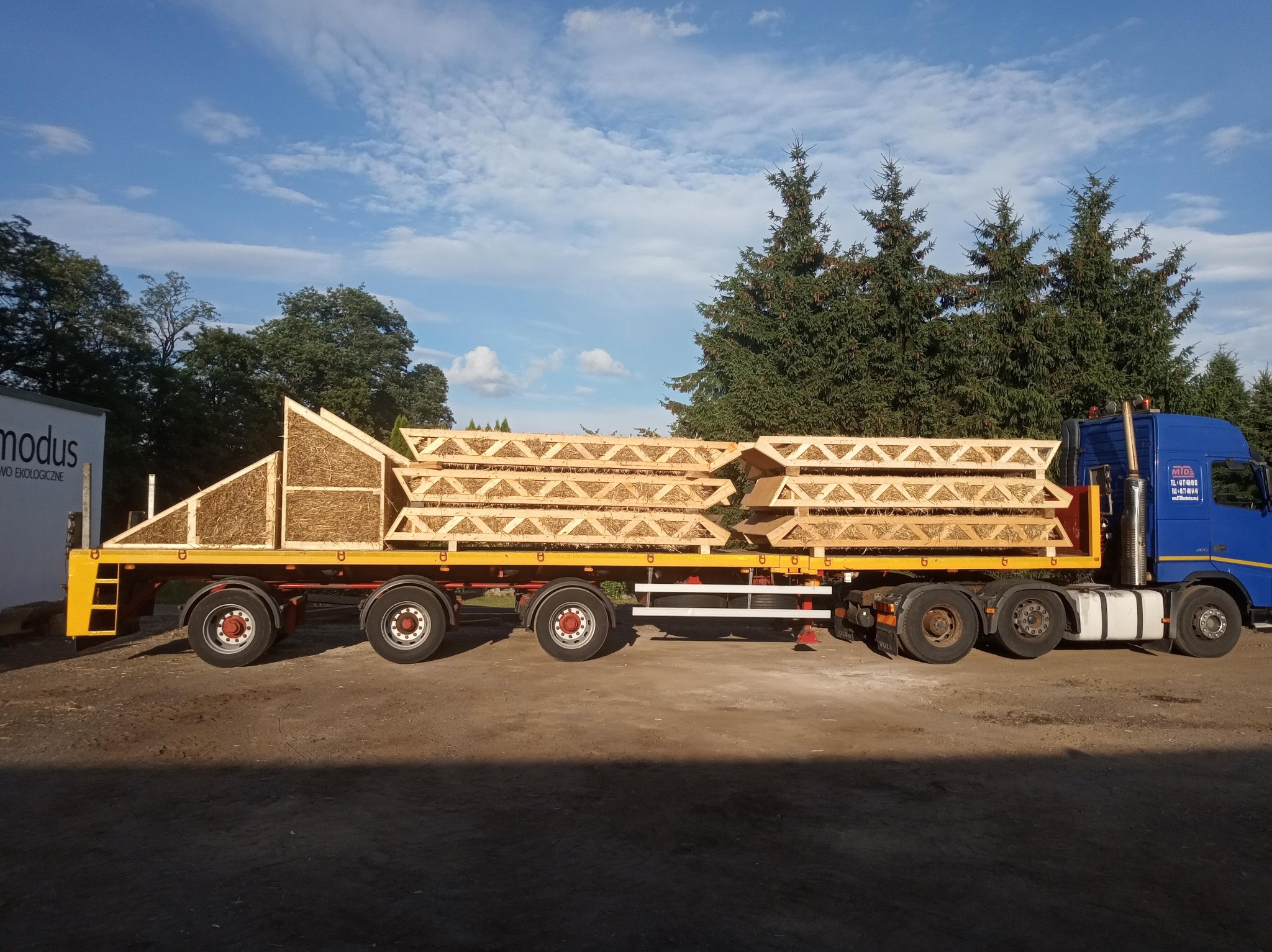Ciężarówka transportuje elementy konstrukcyjne domu szkieletowego wypełnione słomą, na tle zielonych drzew i błękitnego nieba.