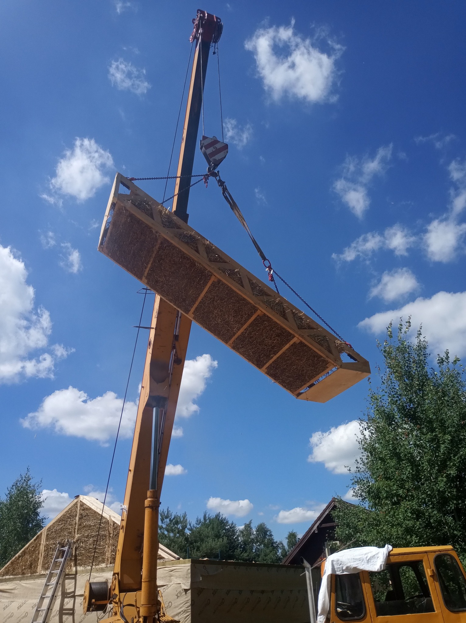 Żuraw przenosi prefabrykowany moduł ze słomy na budowę domu w słoneczny dzień, widoczne fragmenty elewacji budynku w tle.